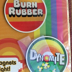 May include: Two round magnets with colourful designs. The first magnet is red with yellow stripes and the text "BURN RUBBER". The second magnet is green with yellow and orange stripes and the text "DYNOMITE".