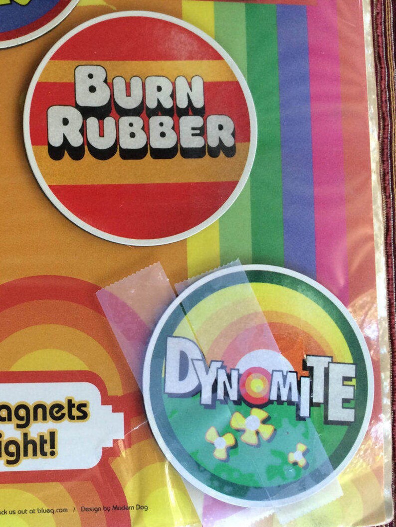 May include: Two round magnets with colourful designs. The first magnet is red with yellow stripes and the text "BURN RUBBER". The second magnet is green with yellow and orange stripes and the text "DYNOMITE".