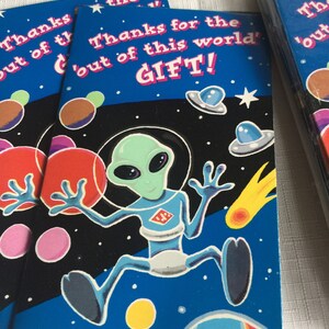 May include: A blue thank you card with a cartoon alien in a blue and white spacesuit. The card has a black background with colorful planets and stars. The text on the card reads "Thanks for the 'out of this world' GIFT!"