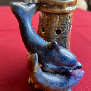 May include: A ceramic lighthouse figurine with two blue dolphins wrapped around the base. The lighthouse is decorated with brown and blue glaze.