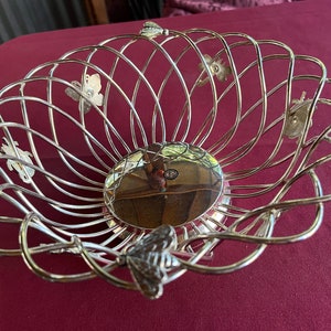 Op de afbeelding: Zilveren draad fruitmand met een decoratief vlinderontwerp. De mand is gemaakt van metaal en heeft een glanzende afwerking.