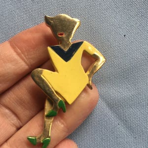 Puede incluir: Un broche dorado que representa a una mujer estilizada con un vestido amarillo y un cuello negro y azul. La mujer est&aacute; sentada con las piernas cruzadas y lleva zapatos verdes.