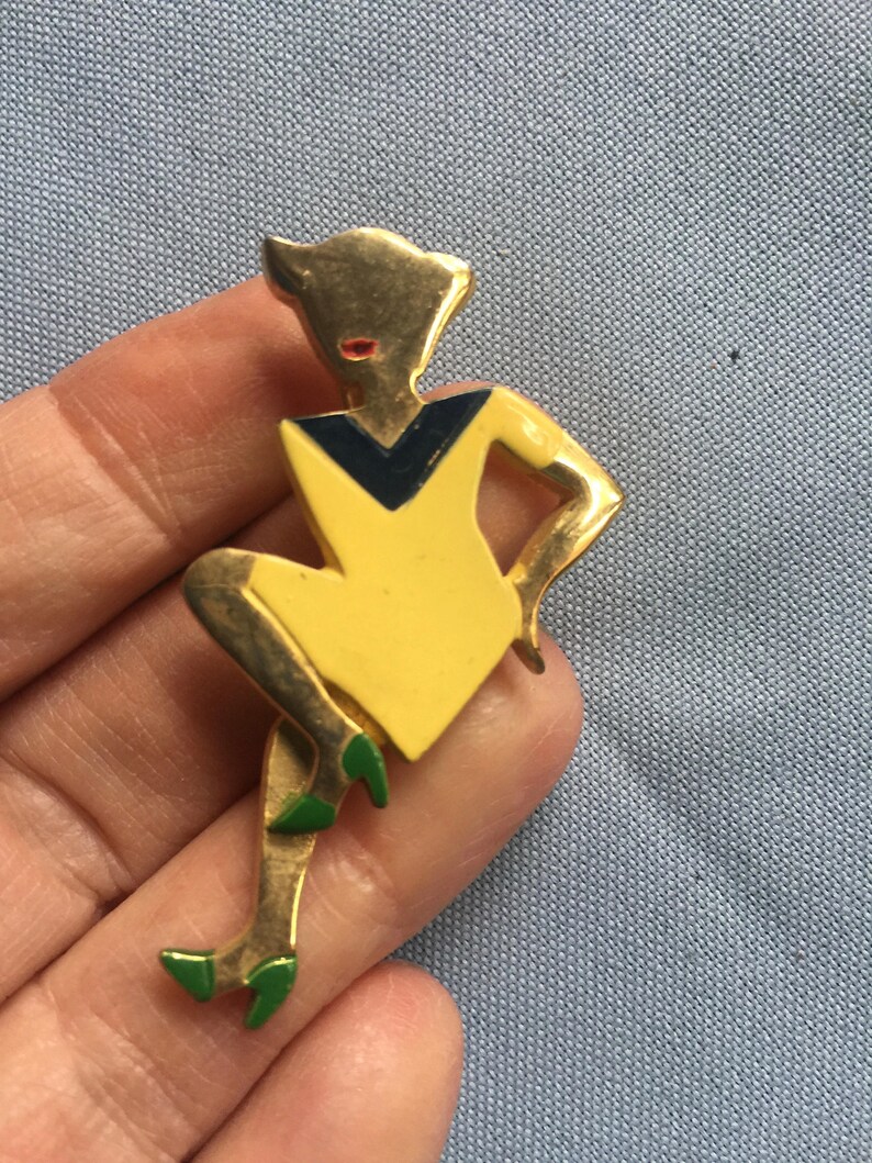 Puede incluir: Un broche dorado que representa a una mujer estilizada con un vestido amarillo y un cuello negro y azul. La mujer est&aacute; sentada con las piernas cruzadas y lleva zapatos verdes.
