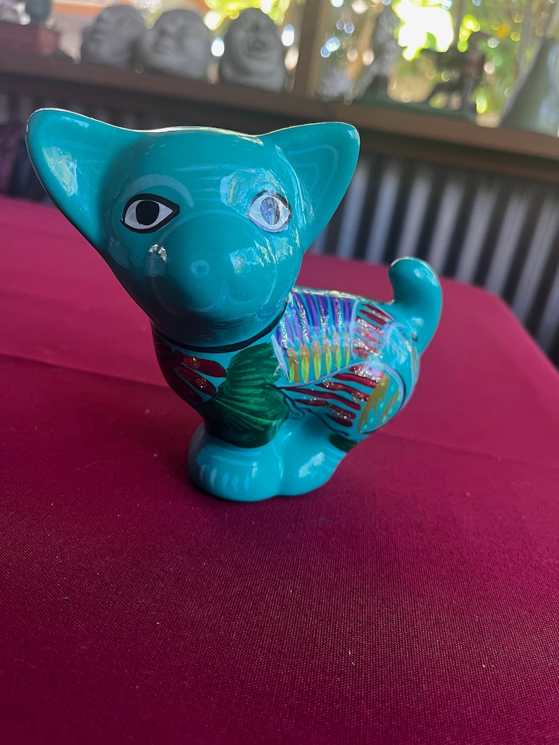 May include: A teal blue ceramic figurine of a dog with colorful painted details. The dog has black eyes and a white nose.