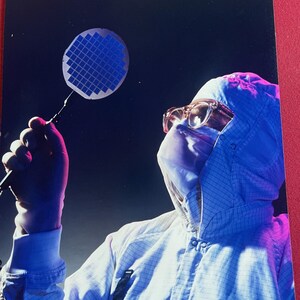 May include: A person in a white grid-patterned cleanroom suit, hood, and protective glasses holds a circular blue silicon wafer with a grid of square chips. The scene is illuminated with blue and magenta lighting against a dark background.