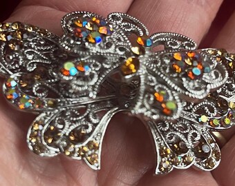 Puede incluir: Una pinza para el pelo de mariposa plateada con pedrería iridiscente en tonos naranja, amarillo y verde.