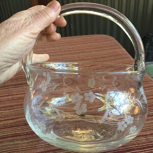 May include: A clear glass bowl with a handle, etched with a floral design. The bowl is sitting on a brown and white patterned surface.