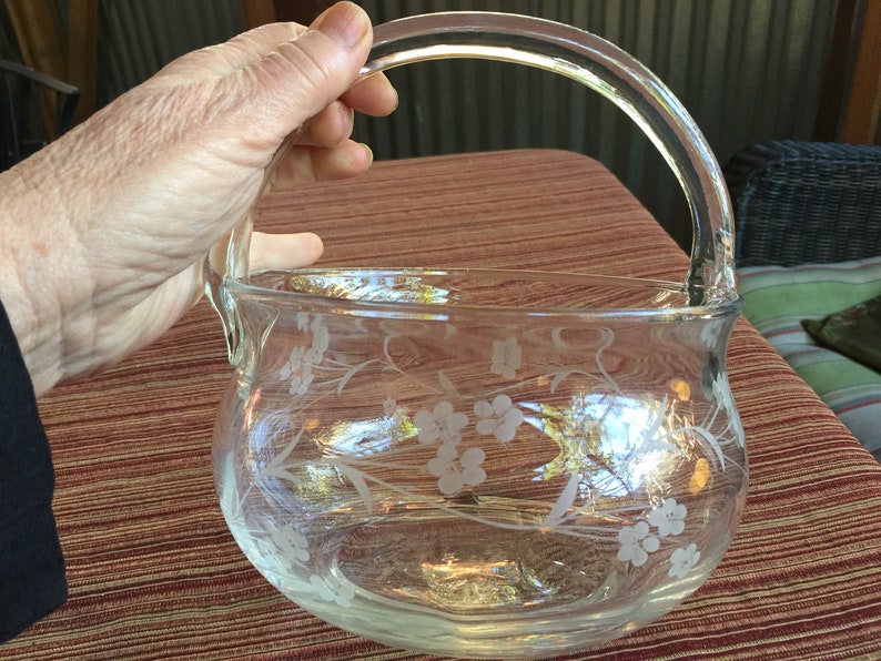 May include: A clear glass bowl with a handle, etched with a floral design. The bowl is sitting on a brown and white patterned surface.