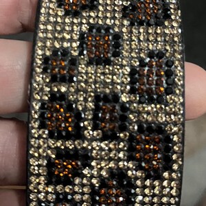 May include: A rectangular bracelet with a leopard print design. The bracelet is covered in small, sparkling rhinestones in shades of brown, black, and gold. The overall effect is a glamorous, eye-catching accessory.