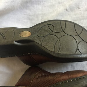 May include: Brown leather clogs with a black rubber sole. The sole has a textured pattern of circles.