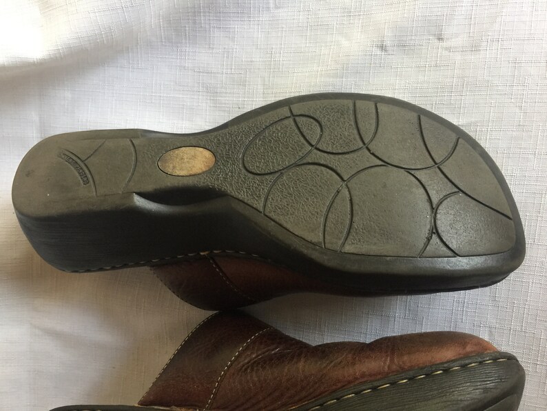 May include: Brown leather clogs with a black rubber sole. The sole has a textured pattern of circles.