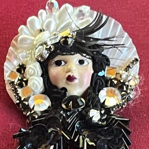 May include: A decorative brooch featuring a porcelain doll face with dark hair and a red lip. The doll is adorned with a white and black hat, embellished with sequins, crystals, and faux pearls. The brooch has a vintage aesthetic.