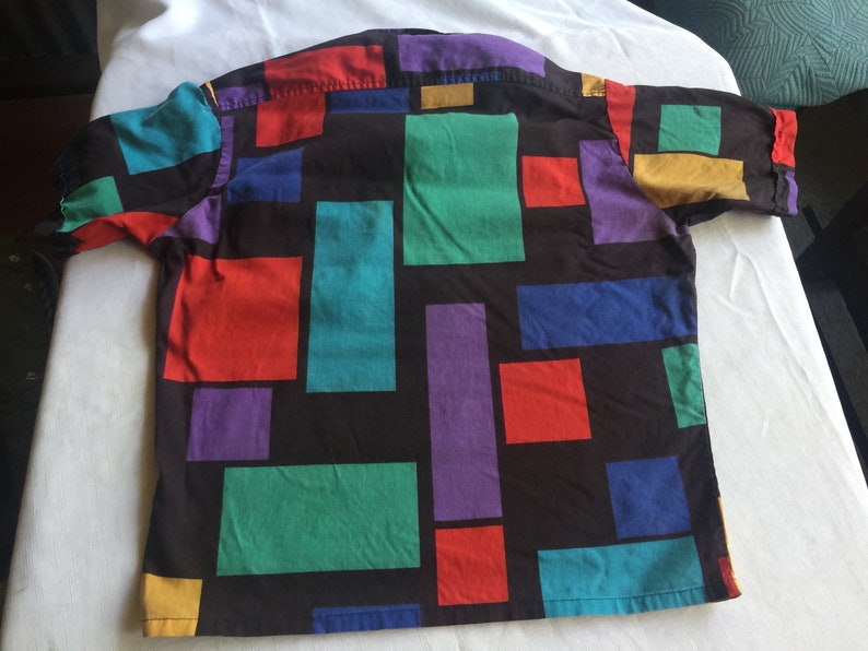 K&ouml;nnte beinhalten: Ein kurz&auml;rmeliges Hemd mit Kn&ouml;pfen und einem geometrischen Muster in Rot-, Blau-, Gr&uuml;n-, Lila- und Gelbt&ouml;nen auf schwarzem Hintergrund.