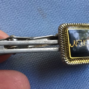 以下が含まれることがあります： 黒い正方形のエナメルの上部に銀色のネクタイピン。エナメルの上部は金色の縁取りがされており、金色の文字で「NEW」と書かれています。