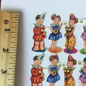 Peut inclure: Un ensemble de huit poup&eacute;es en papier vintage, chacune v&ecirc;tue d'un costume diff&eacute;rent, y compris un clown, une fille avec une guitare et une fille avec un violon. Les poup&eacute;es sont imprim&eacute;es sur du papier &eacute;pais et ont un design color&eacute;. Les poup&eacute;es sont num&eacute;rot&eacute;es 869 et indiquent "Fabriqu&eacute; en Angleterre".