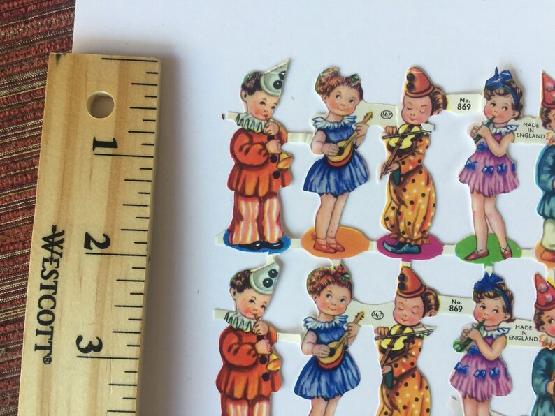 Peut inclure: Un ensemble de huit poup&eacute;es en papier vintage, chacune v&ecirc;tue d'un costume diff&eacute;rent, y compris un clown, une fille avec une guitare et une fille avec un violon. Les poup&eacute;es sont imprim&eacute;es sur du papier &eacute;pais et ont un design color&eacute;. Les poup&eacute;es sont num&eacute;rot&eacute;es 869 et indiquent "Fabriqu&eacute; en Angleterre".