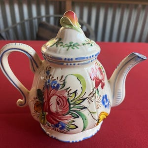 以下が含まれることがあります： 花柄の白い陶器製のティーポット。ティーポットの蓋にはピンクと緑の花が飾られています。ティーポットは青、赤、緑、黄色の花で飾られています。