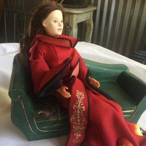 May include: A doll wearing a red and black robe sits on a green miniature couch. The doll has long brown hair and is looking to the right.