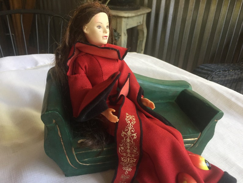 May include: A doll wearing a red and black robe sits on a green miniature couch. The doll has long brown hair and is looking to the right.