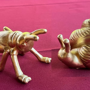 May include: Two gold-painted figurines, a grasshopper and a snail, on a red background. The grasshopper is standing on all six legs, with its head tilted up. The snail is curled up in its shell, with its head and tentacles extended.