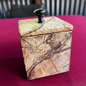 May include: A square, brown and beige marble box with a black knob on the lid. The box is sitting on a red surface.