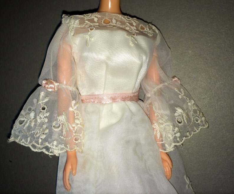 Puede incluir: Un vestido blanco de encaje con una cintura de cinta rosa y mangas acampanadas. El vestido est&aacute; hecho de una tela transparente y tiene un delicado patr&oacute;n floral.