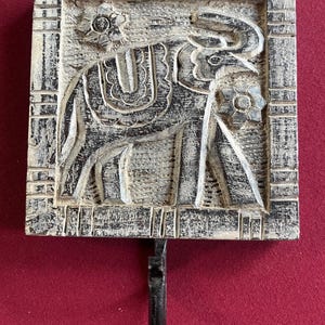 May include: A square, distressed-look wall hook with a carved elephant design. The elephant is adorned with floral accents and a saddle. The hook is black, and the frame is off-white with black distressing. The hook is likely made of wood.