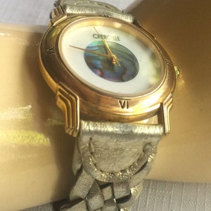 May include: A gold-tone watch with a white face and a braided leather band. The watch face features a round, iridescent abalone shell in the center and the word "CHerokee" printed at the top.