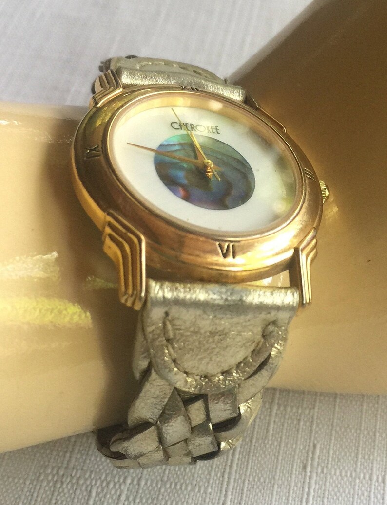 May include: A gold-tone watch with a white face and a braided leather band. The watch face features a round, iridescent abalone shell in the center and the word "CHerokee" printed at the top.