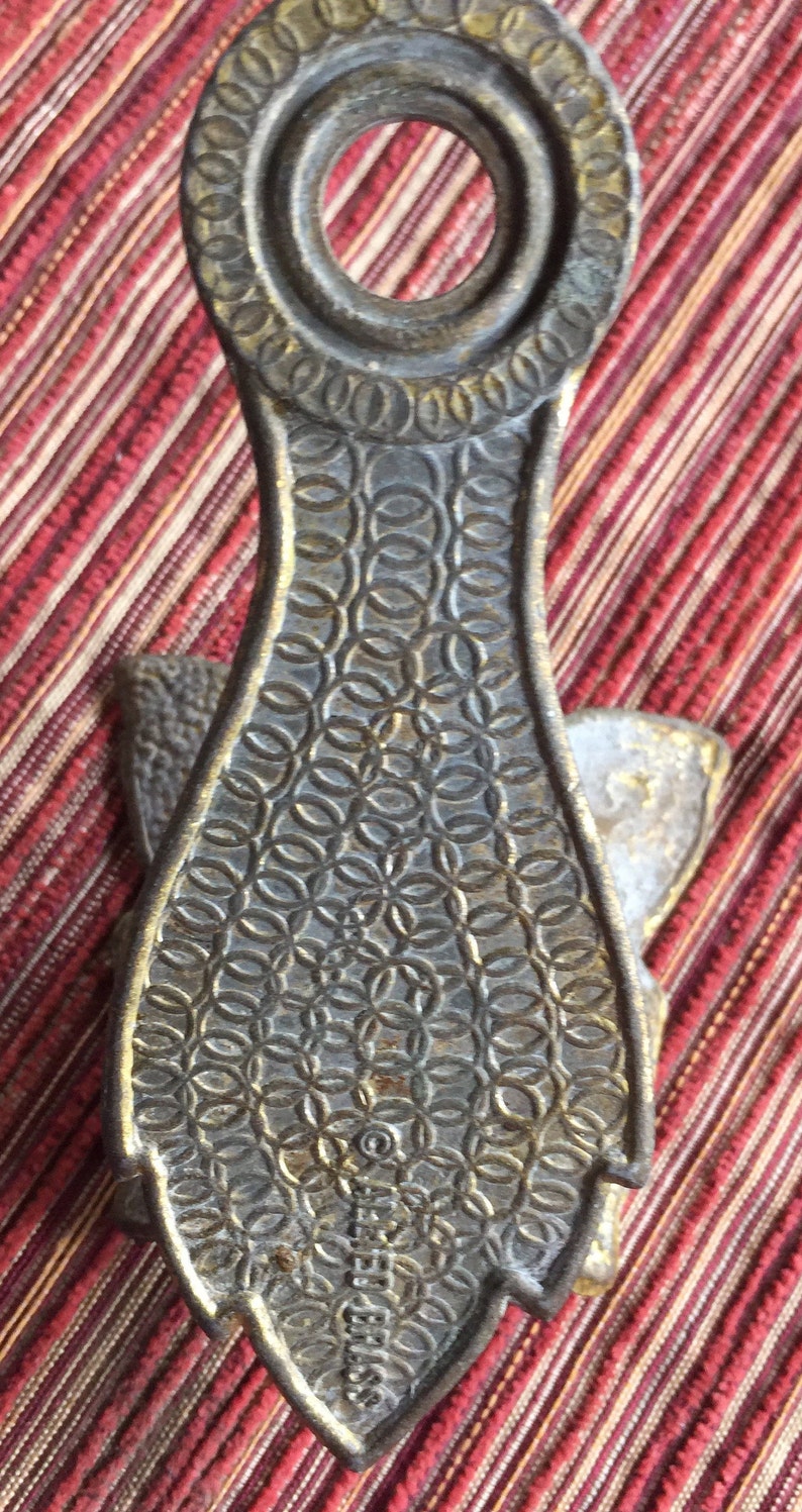 May include: A vintage brass bottle opener with a decorative design. The opener features a circular handle and a patterned body with a shield-shaped end. The text "SEIFERT BRASS" is visible on the bottom.