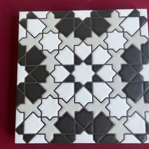 May include: Square tile with a repeating geometric pattern in black, white, and gray. The design features eight-pointed star shapes and diamond-like forms, creating a symmetrical and visually appealing surface.