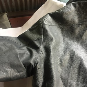 Pu&ograve; includere: Una giacca di pelle grigio scuro con una superficie testurizzata. La giacca ha una chiusura a zip e una tasca a lembo con bottone sul lato sinistro.