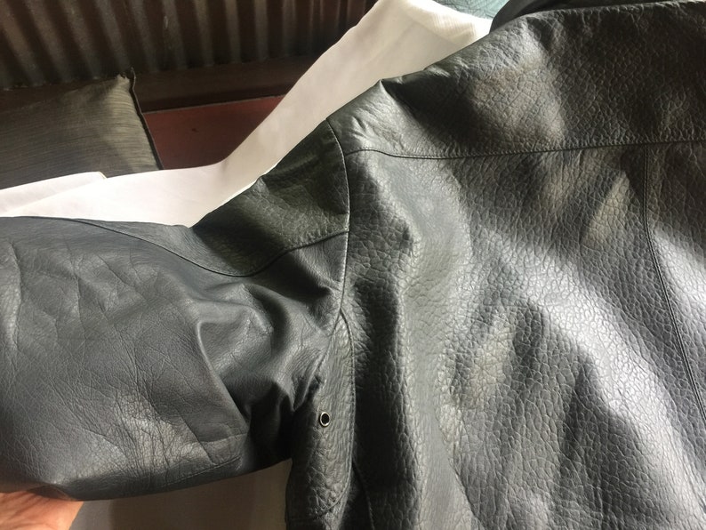 Pu&ograve; includere: Una giacca di pelle grigio scuro con una superficie testurizzata. La giacca ha una chiusura a zip e una tasca a lembo con bottone sul lato sinistro.