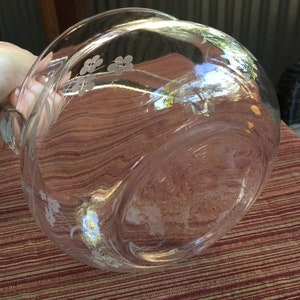 May include: A clear glass serving bowl with a floral design etched into the side. The bowl has a handle and is sitting on a red and brown patterned surface.