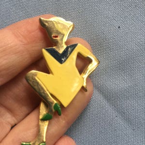 Puede incluir: Un broche dorado que representa a una mujer estilizada con un vestido amarillo con un escote negro y azul. La mujer lleva zapatos verdes y tiene la cara roja.