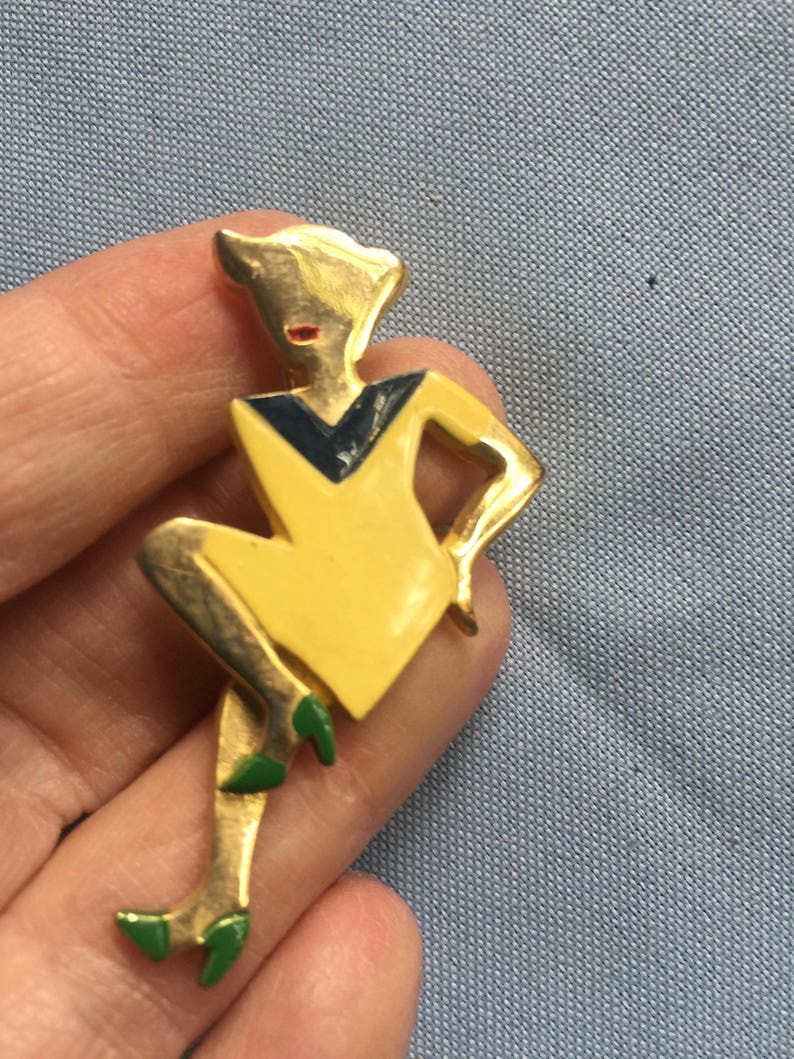Puede incluir: Un broche dorado que representa a una mujer estilizada con un vestido amarillo con un escote negro y azul. La mujer lleva zapatos verdes y tiene la cara roja.