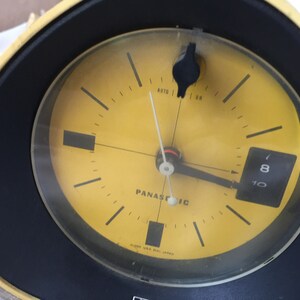 May include: A yellow analog alarm clock with black hands and a black bezel. The clock face has the word "Panasonic" printed on it and the text "Clock U.S.A. Mfd. Japan". The clock has a small black knob on the right side of the face labeled "Auto" and "On".