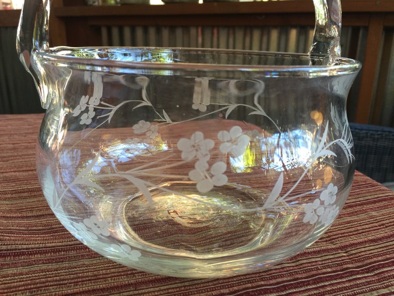 May include: A clear glass bowl with a floral design etched into the side. The bowl has a handle and is perfect for serving snacks or fruit.