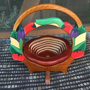 Op de afbeelding: Een houten fruitmand met een handvat, beschilderd met een kleurrijk ontwerp van groenten, waaronder tomaten, maïs en aubergines. De mand is gemaakt van hout en heeft een bruine vlek.