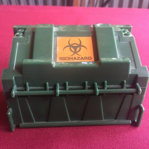 May include: A green plastic toy dumpster with a hinged lid and a yellow biohazard sign on the top. The sign has a black biohazard symbol and the word "BIOHAZARD" printed in black.