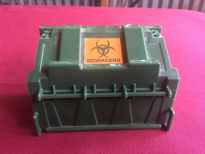 May include: A green plastic toy dumpster with a hinged lid and a yellow biohazard sign on the top. The sign has a black biohazard symbol and the word "BIOHAZARD" printed in black.