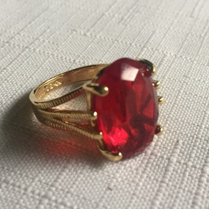 May include: A gold ring with a large, red, faceted gemstone. The ring has a simple band with a delicate design.