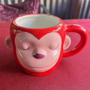 May include: Red ceramic coffee mug with a monkey face design. The monkey has closed eyes and a pink nose.