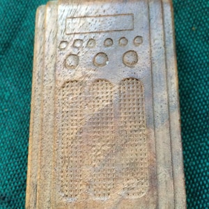 May include: A wooden carving of a vintage radio with a textured speaker grill and a row of round knobs. The carving is painted in shades of brown and beige.
