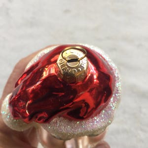 以下が含まれることがあります： 赤く光沢のある上部と白くキラキラしたベースのクリスマスオーナメント。オーナメントには「Poland」の文字がプリントされた金色の金属キャップが付いています。オーナメントは手で持たれています。