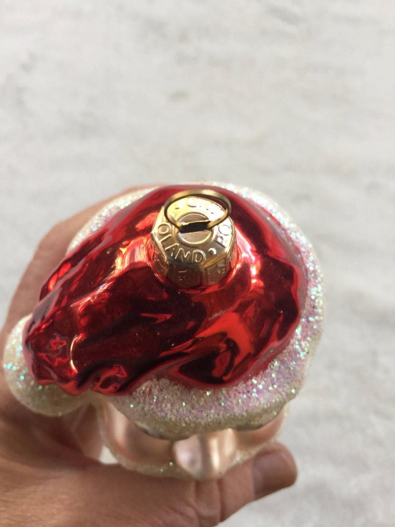 以下が含まれることがあります： 赤く光沢のある上部と白くキラキラしたベースのクリスマスオーナメント。オーナメントには「Poland」の文字がプリントされた金色の金属キャップが付いています。オーナメントは手で持たれています。