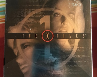 May include: The X-Files: The Complete First Season DVD Collector's Edition. The cover features a black and white image of two people's faces, with the number 1 and the show's title in white. The DVD case is on a colorful striped surface.