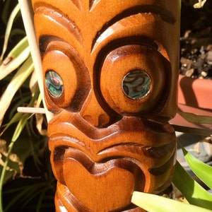 Könnte beinhalten: Eine Holz-Tiki-Statue mit einem geschnitzten Gesicht und eingelegten Augen aus Abalone-Muscheln. Die Statue ist braun und hat ein herzförmiges Design auf der Brust.