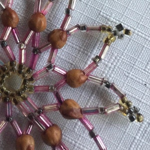 Pode incluir: Um close-up de um ornamento de floco de neve de contas rosa e prata com contas de madeira marrom. O ornamento &eacute; feito com contas pequenas e iridescentes e tem um design delicado e intrincado.