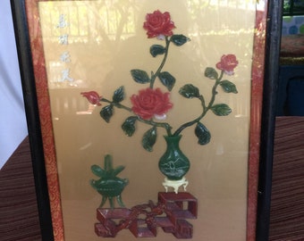 May include: A framed artwork featuring a jade carving of a rose bush with red flowers in a green vase. The artwork is set against a gold background with Chinese characters.